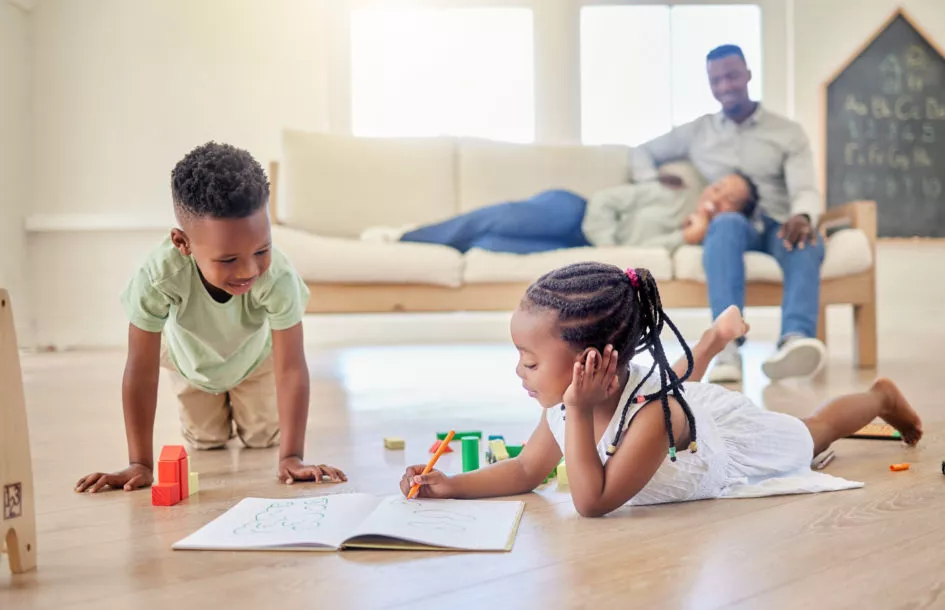 teaching gratitude to children: Little kids lying on the floor with toys and colouring in a book. Small african american brother and sister playing together at home while parents sit in the background.