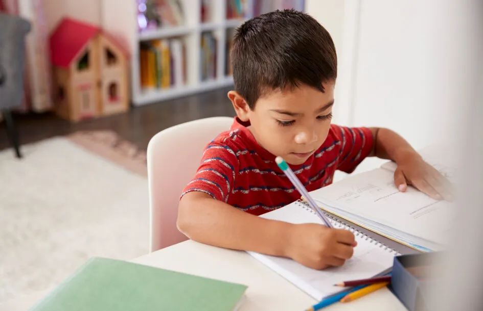 Young Boy Sitting At Desk In Bedroom Doing Homework with a pencil, practicing gross motor skills for children