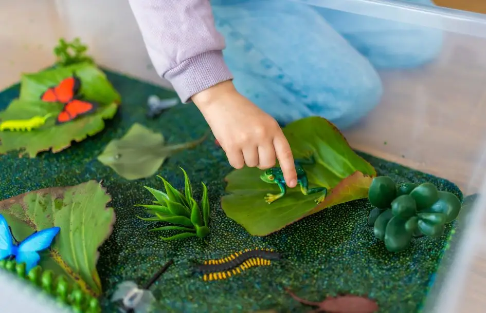 Little girl playing in handmade swamp of green-dyed chia seeds with insect, fish and plant models for spring sensory bins for kids