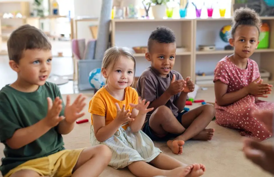Group of small nursery school children sitting on floor indoors in classroom, clapping. for rhyming and leter games, early reading activities for kids